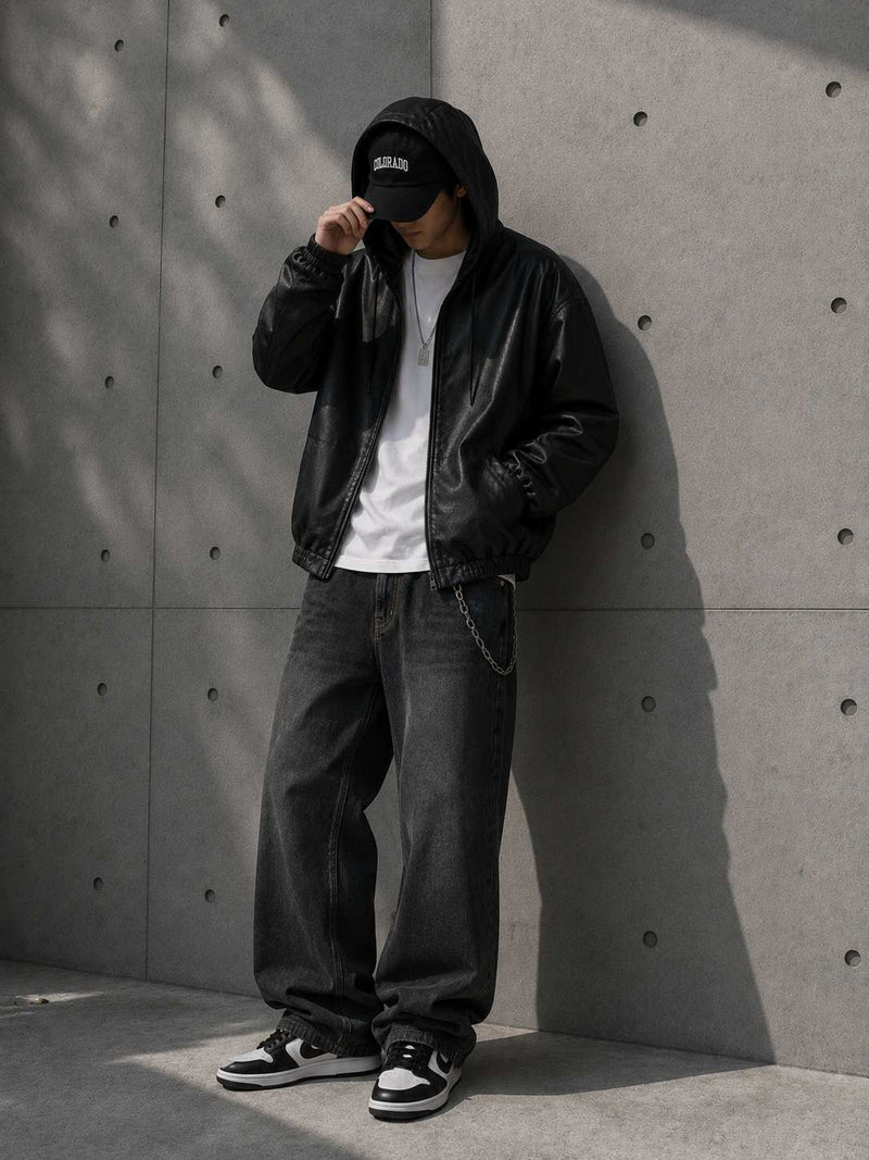 Person wearing a black leather jacket, white shirt, and black pants against a concrete wall.
