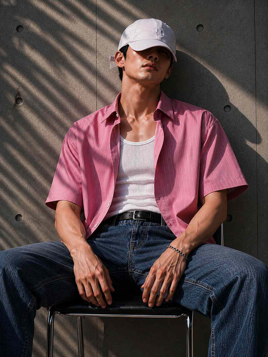 Person wearing a pink shirt, white cap, and blue jeans sitting against a textured wall.
