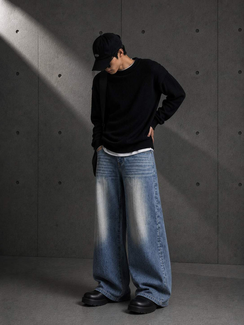 Person wearing a black sweater, blue jeans, and a black cap against a gray concrete wall.