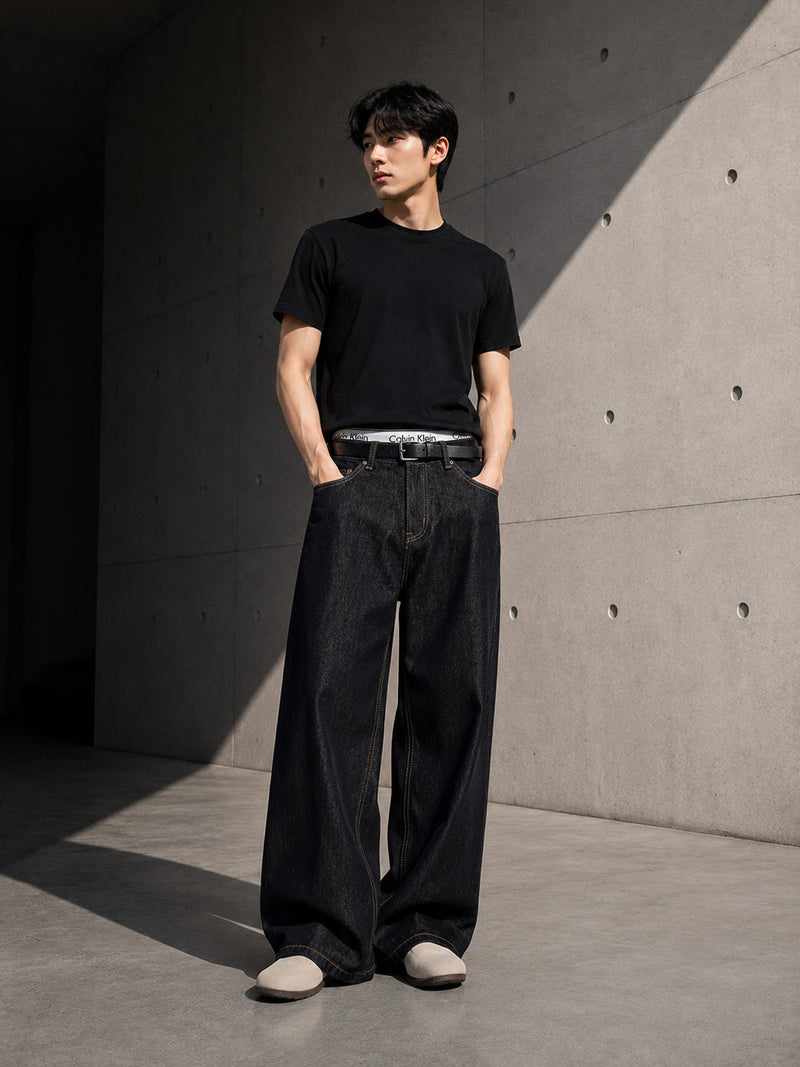 Person wearing a black t-shirt and black straight-leg denim jeans standing against a concrete wall.