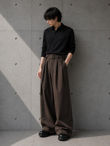 Person wearing a black shirt and brown wide-leg pants against a gray concrete wall.