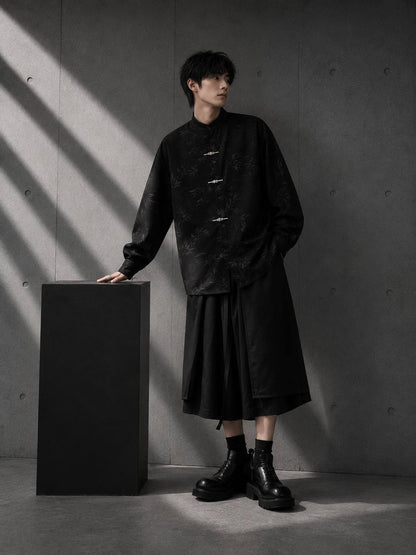 Person wearing a black outfit standing next to a black cube against a gray wall.