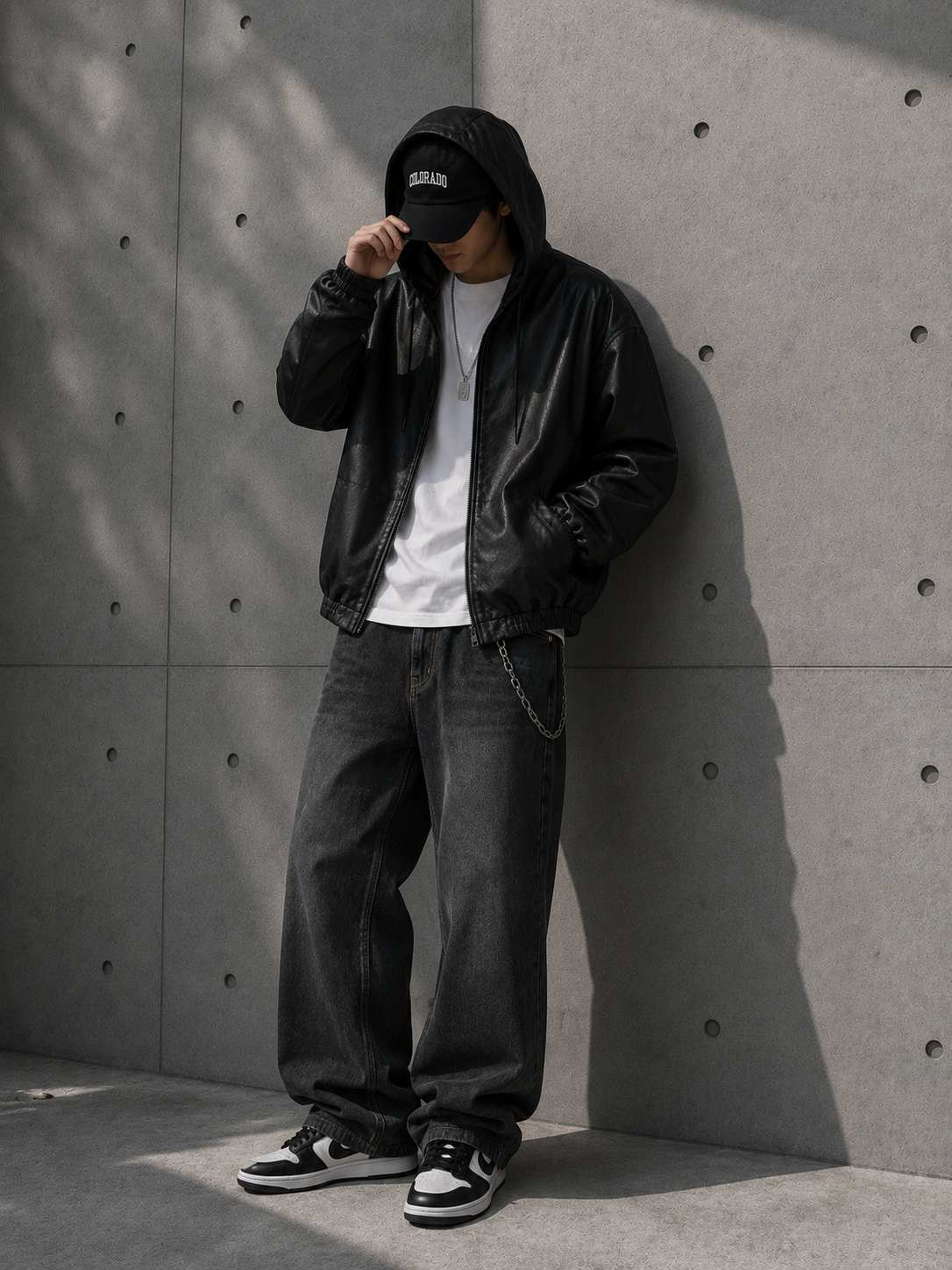 Person wearing a black leather jacket, white shirt, and black pants against a concrete wall.