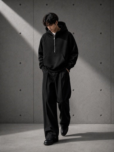 Person wearing a black hoodie and pants standing against a gray concrete wall.