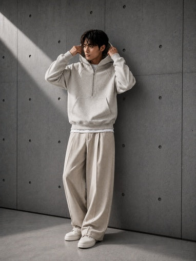 Person wearing a light gray hoodie and beige pants against a textured gray wall.