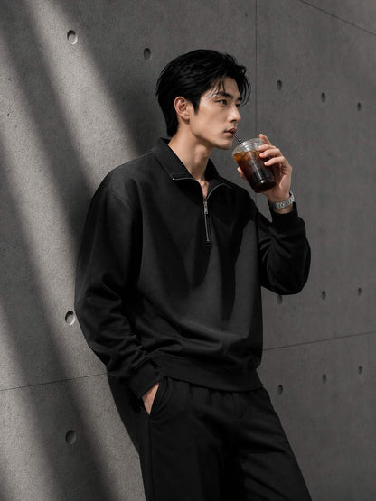 Man in black outfit holding a drink against a gray concrete wall