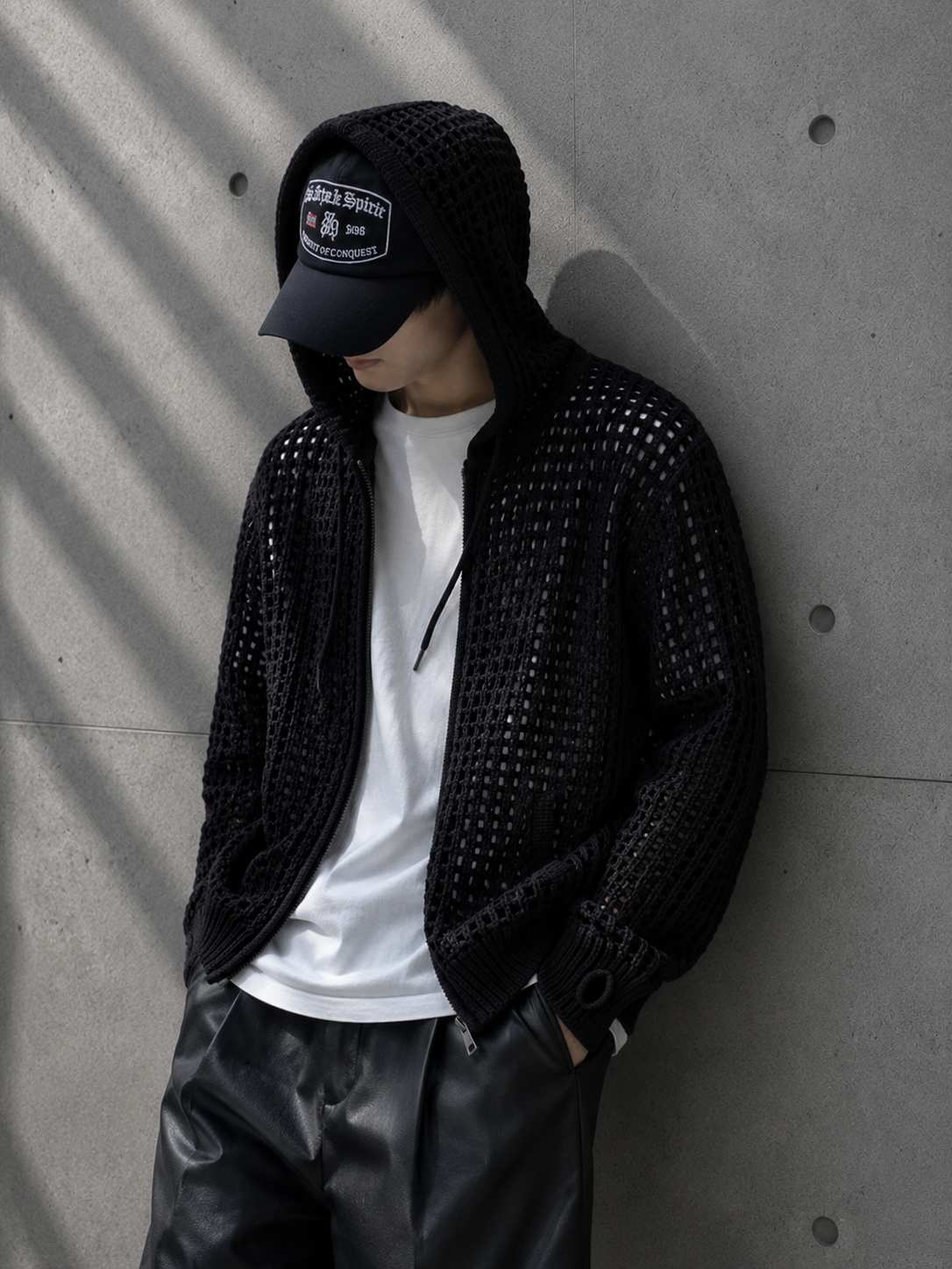 Person wearing a black studded jacket, white t-shirt, and black cap against a concrete wall.