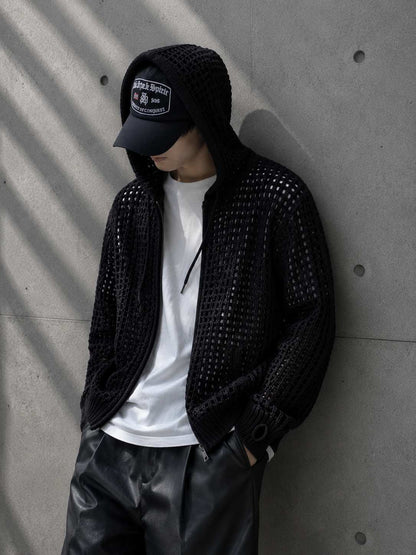 Person wearing a black studded jacket, white t-shirt, and black cap against a concrete wall.