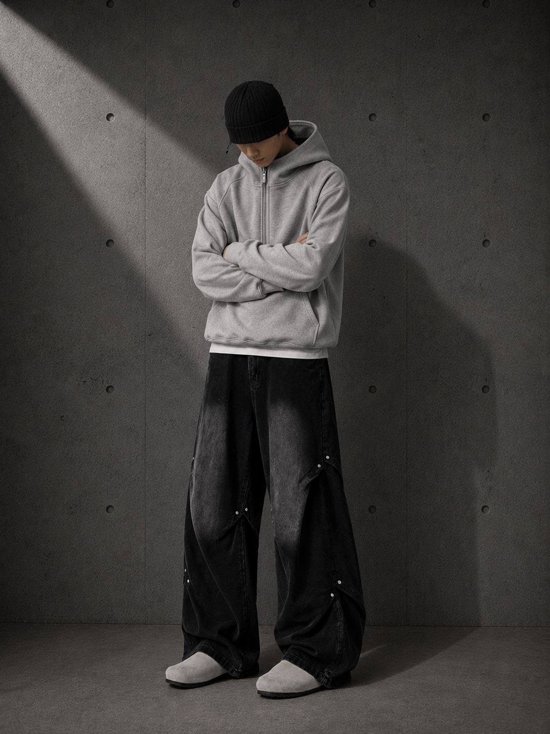 Person wearing a gray hoodie and black pants standing against a textured gray wall.
