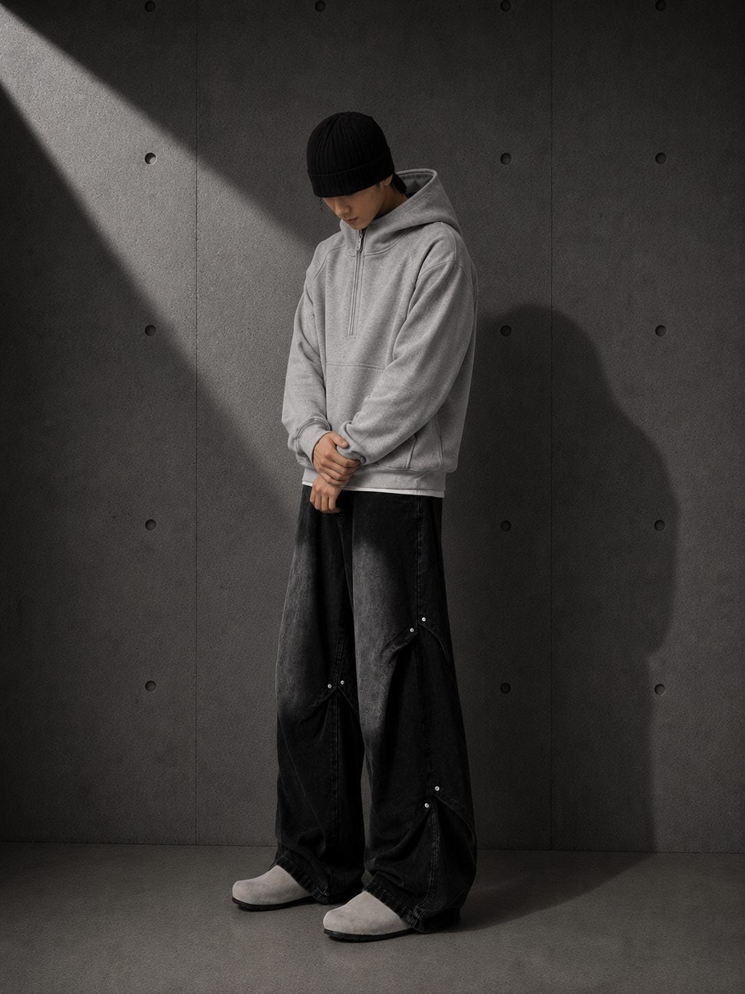 Person wearing a gray hoodie and dark pants against a textured gray wall.