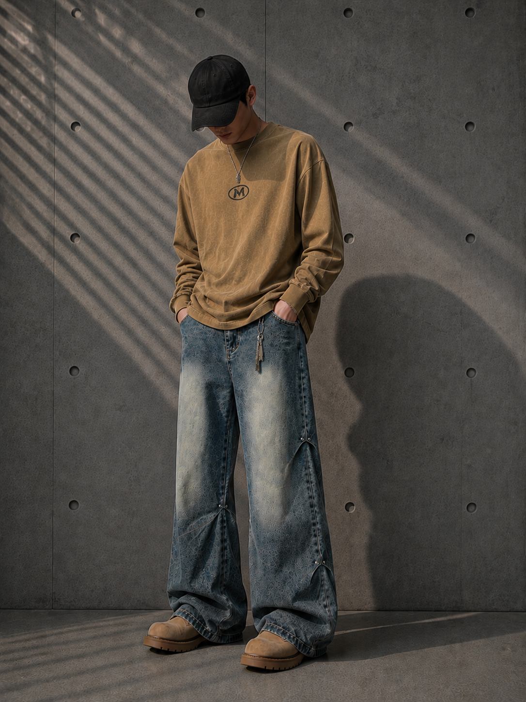 Person wearing a brown long-sleeve shirt, blue jeans, and a black cap against a concrete wall.