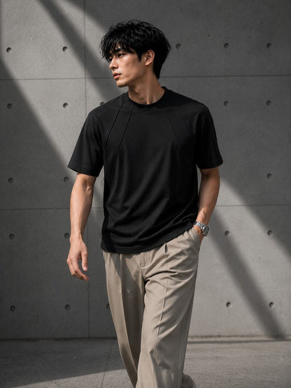 Man wearing a black t-shirt and beige pants against a gray concrete wall.