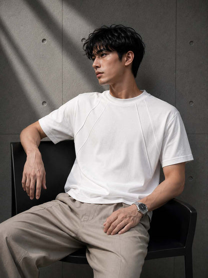 Man wearing a white t-shirt and beige pants sitting against a gray wall.
