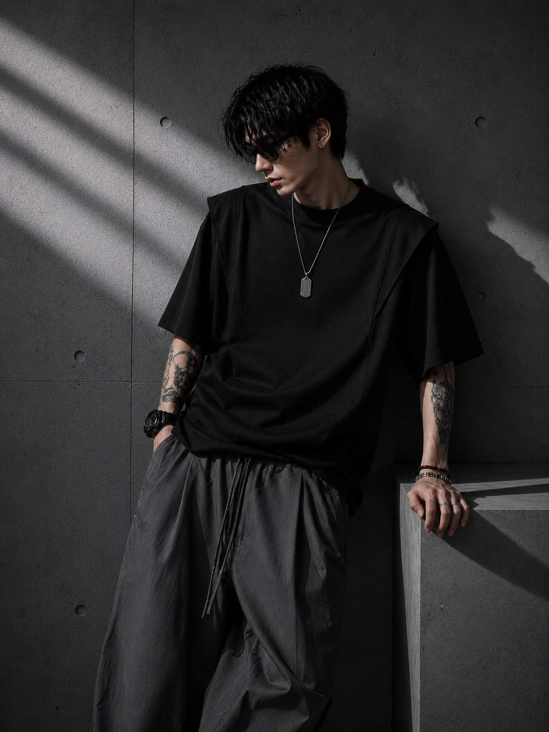 Person wearing a black t-shirt and dark pants against a gray wall with shadows.