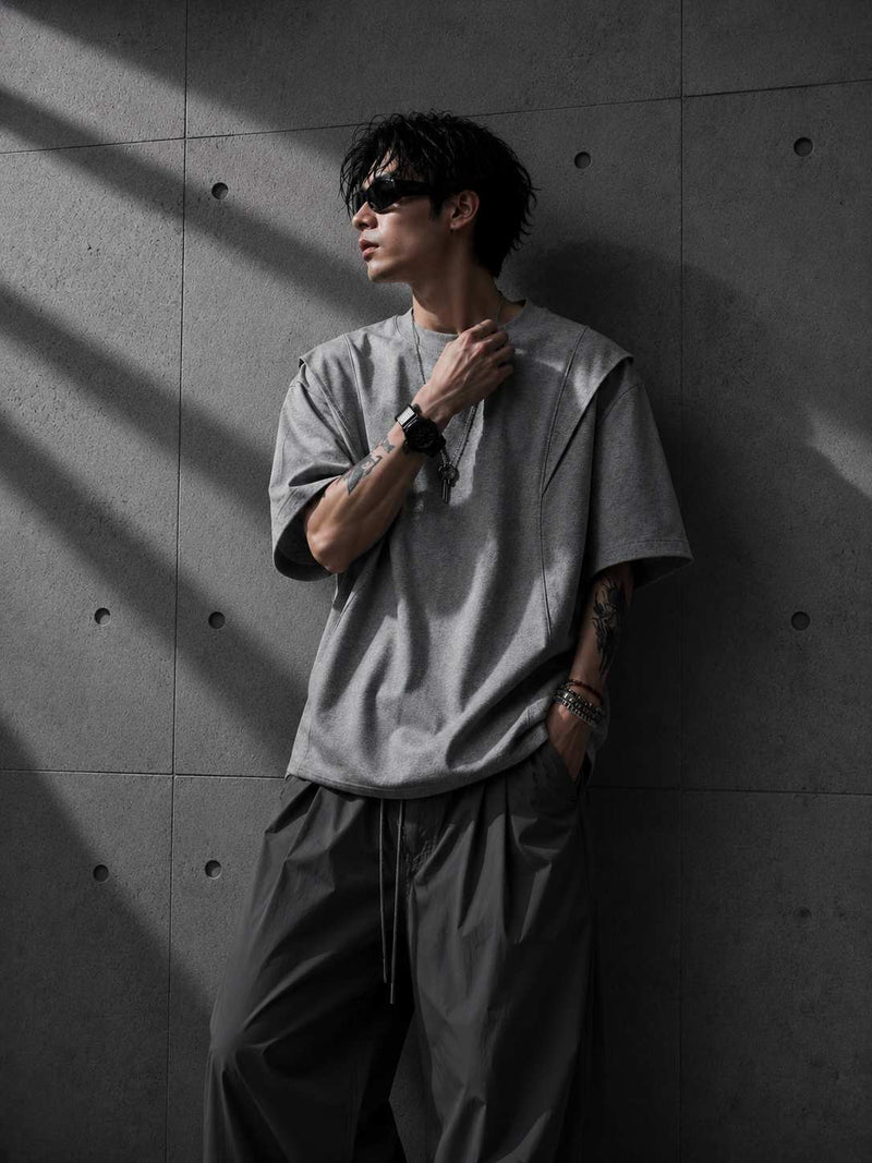 Person wearing a gray oversized t-shirt and dark pants against a textured wall.