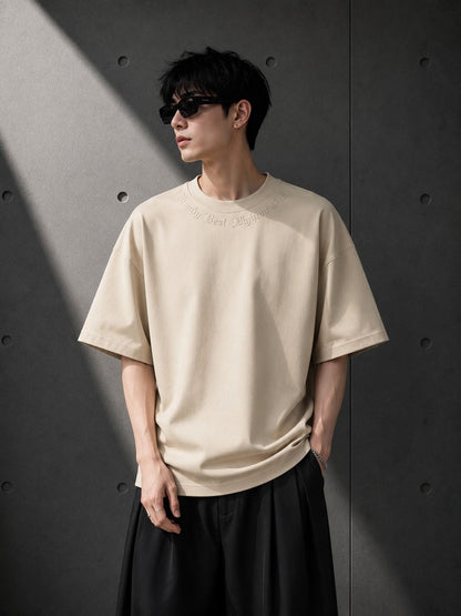 Person wearing a beige t-shirt and black pants against a gray wall.