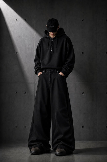 Person wearing a black hoodie and pants standing against a dark concrete wall.