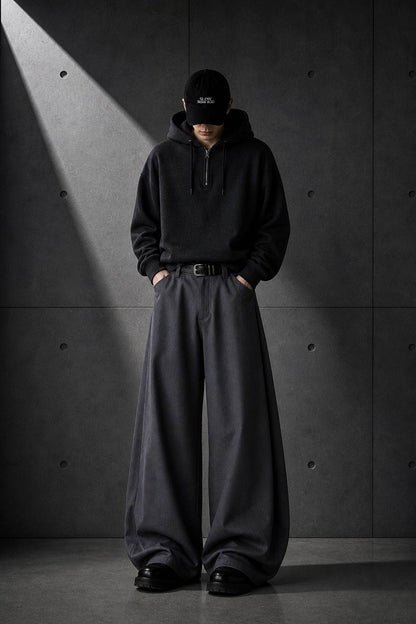 Person wearing a black hoodie and wide-leg pants against a dark concrete wall.