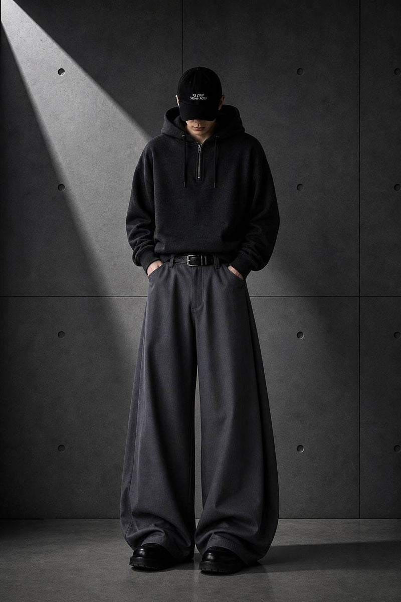 Person wearing a black hoodie and wide-leg pants against a dark concrete wall.