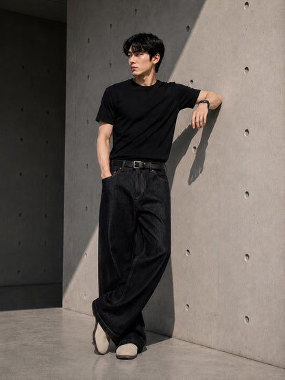 Man wearing a black t-shirt and black straight-leg denim jeans leaning against a concrete wall.