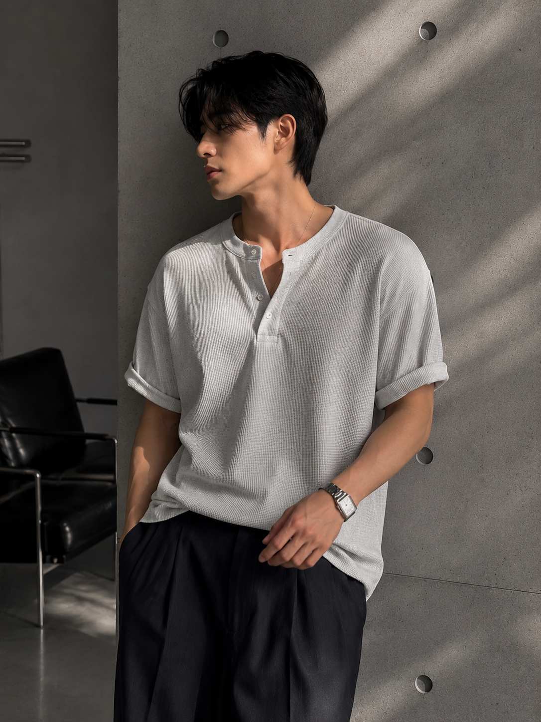 Man wearing a light gray shirt and dark pants against a concrete wall.