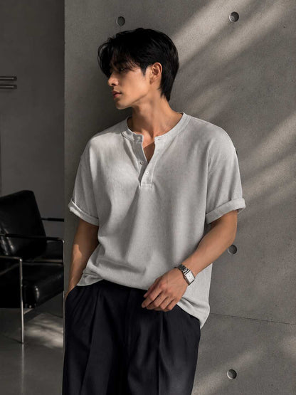 Man wearing a light gray shirt and dark pants against a concrete wall.